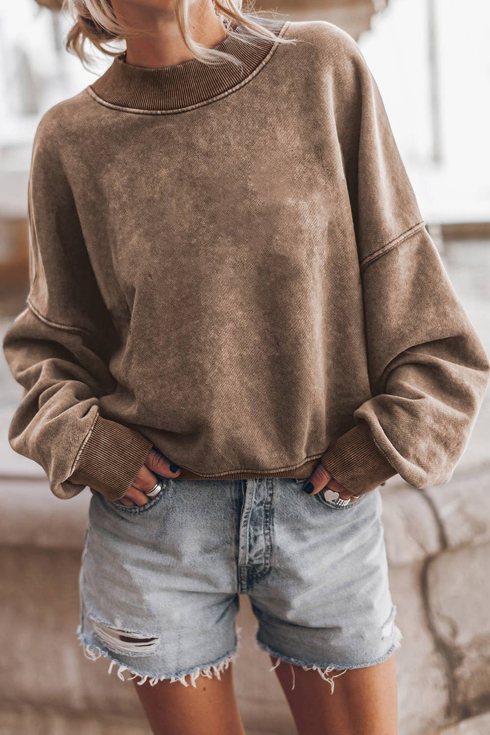 A person wearing an oversized brown washed crewneck sweatshirt and light-wash distressed denim shorts, standing outdoors near a stone wall.