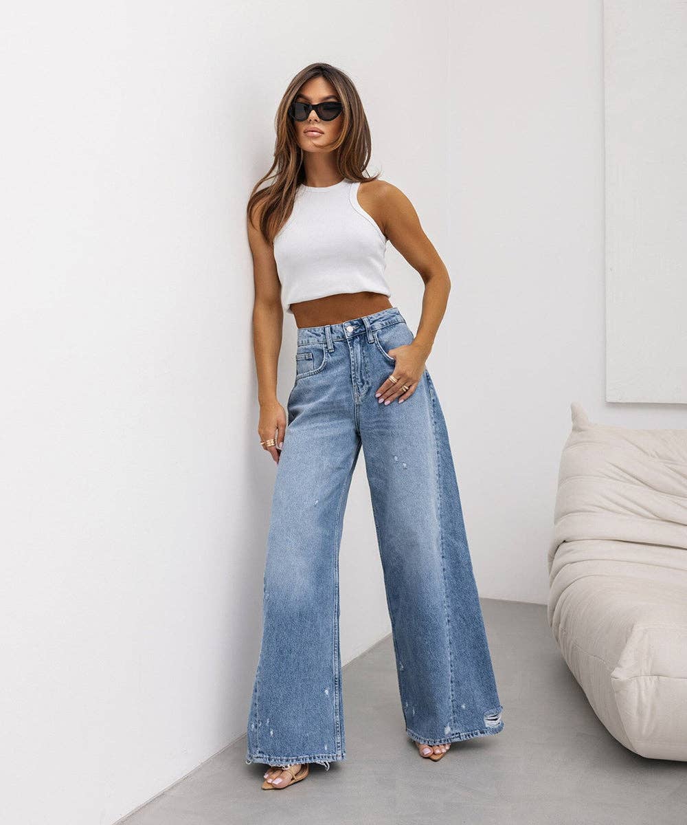A woman modeling a pair of high-waisted, wide-leg light blue denim jeans with a distressed finish, paired with a white crop top and sunglasses in a minimalist room setting.