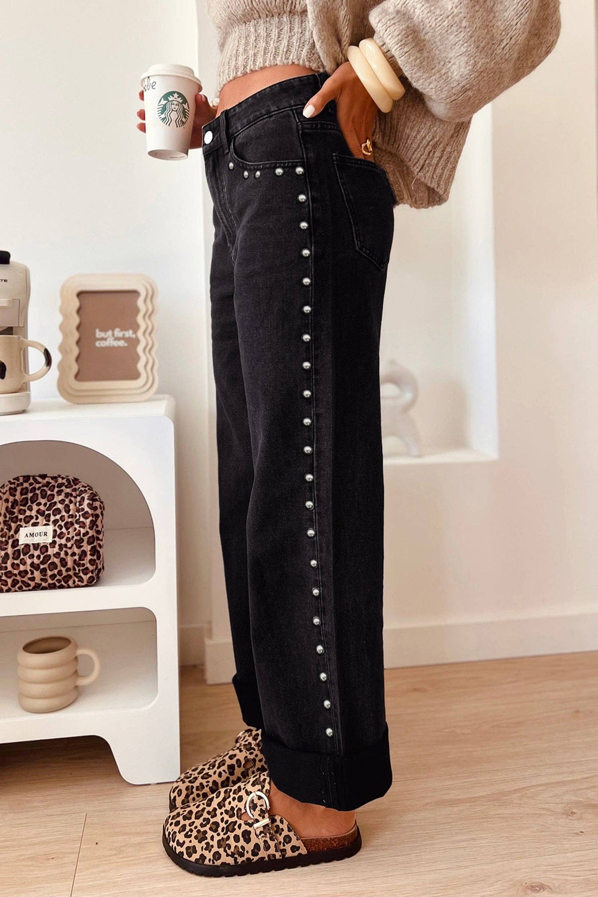 A person wearing black wide-leg jeans with silver stud detailing down the side, paired with leopard-print slippers and a beige knit sweater, standing in a bright indoor room.