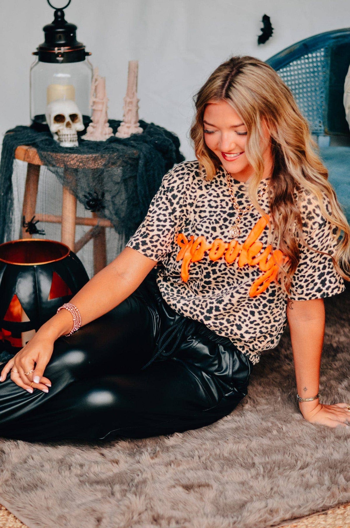 Woman wearing a leopard print shirt with orange text sitting on the floor in a room with Halloween decorations.