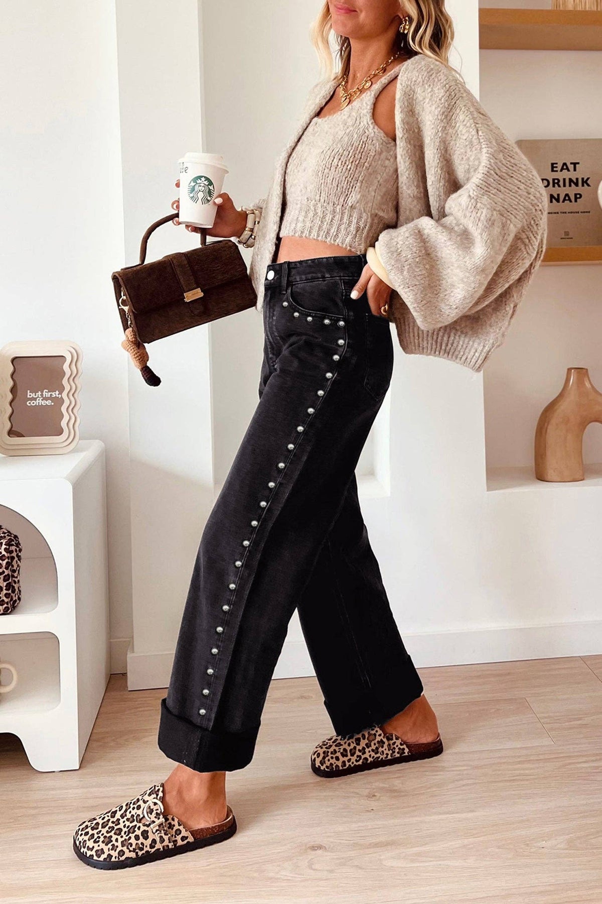 A woman models a pair of black wide-leg jeans featuring silver stud embellishments down the side, styled with leopard-print slippers, a beige knit cardigan, and a brown handbag.