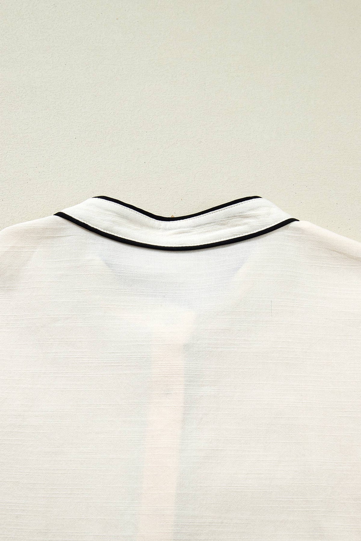 Close-up of a white shirt collar with thin black piping trim, shown against a light neutral background.
