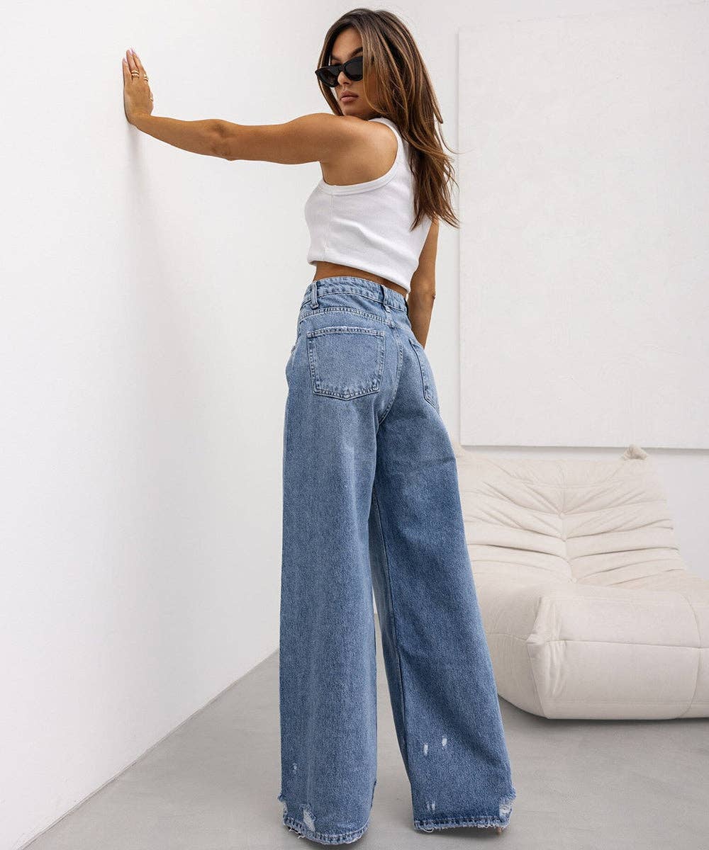 A woman wearing a white cropped tank top and high-waisted, wide-leg blue denim jeans stands in a minimalist room, leaning her hand against a white wall.