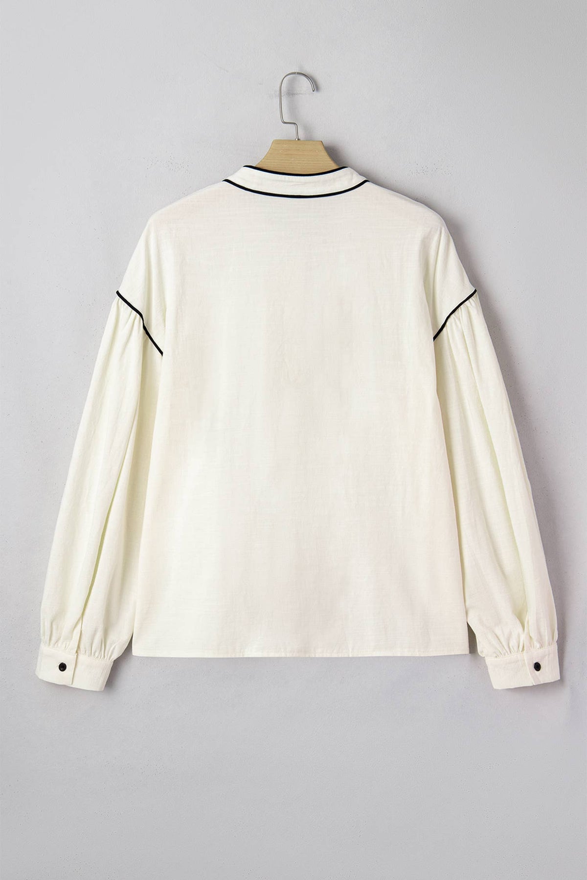 An off-white long-sleeve blouse with black piping trim on the collar and upper sleeves, hanging on a wooden hanger against a neutral gray background.