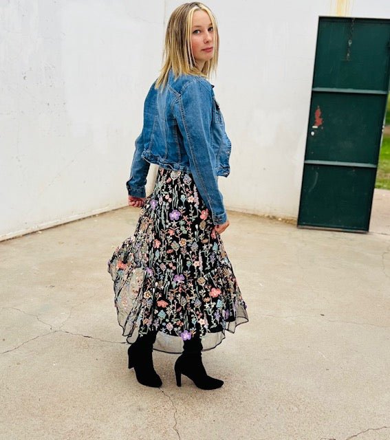 Person wearing a denim jacket and floral skirt standing against a white wall.