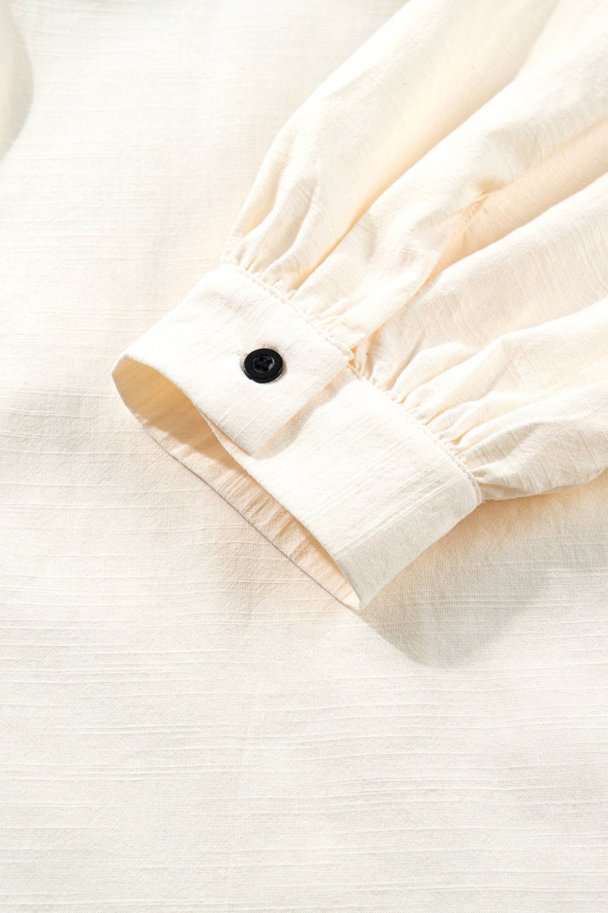 Close-up of a cream-colored fabric garment sleeve with a gathered cuff and a single black button, laid flat on a matching textured fabric background.