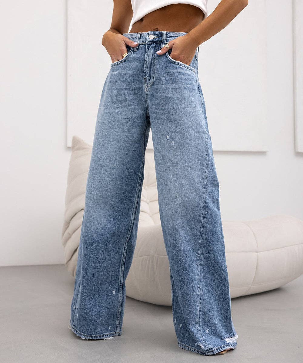 A person wearing light blue wide-leg denim jeans with a faded wash and subtle distressed details, standing in a bright indoor room with a white wall and a beige cushion in the background.
