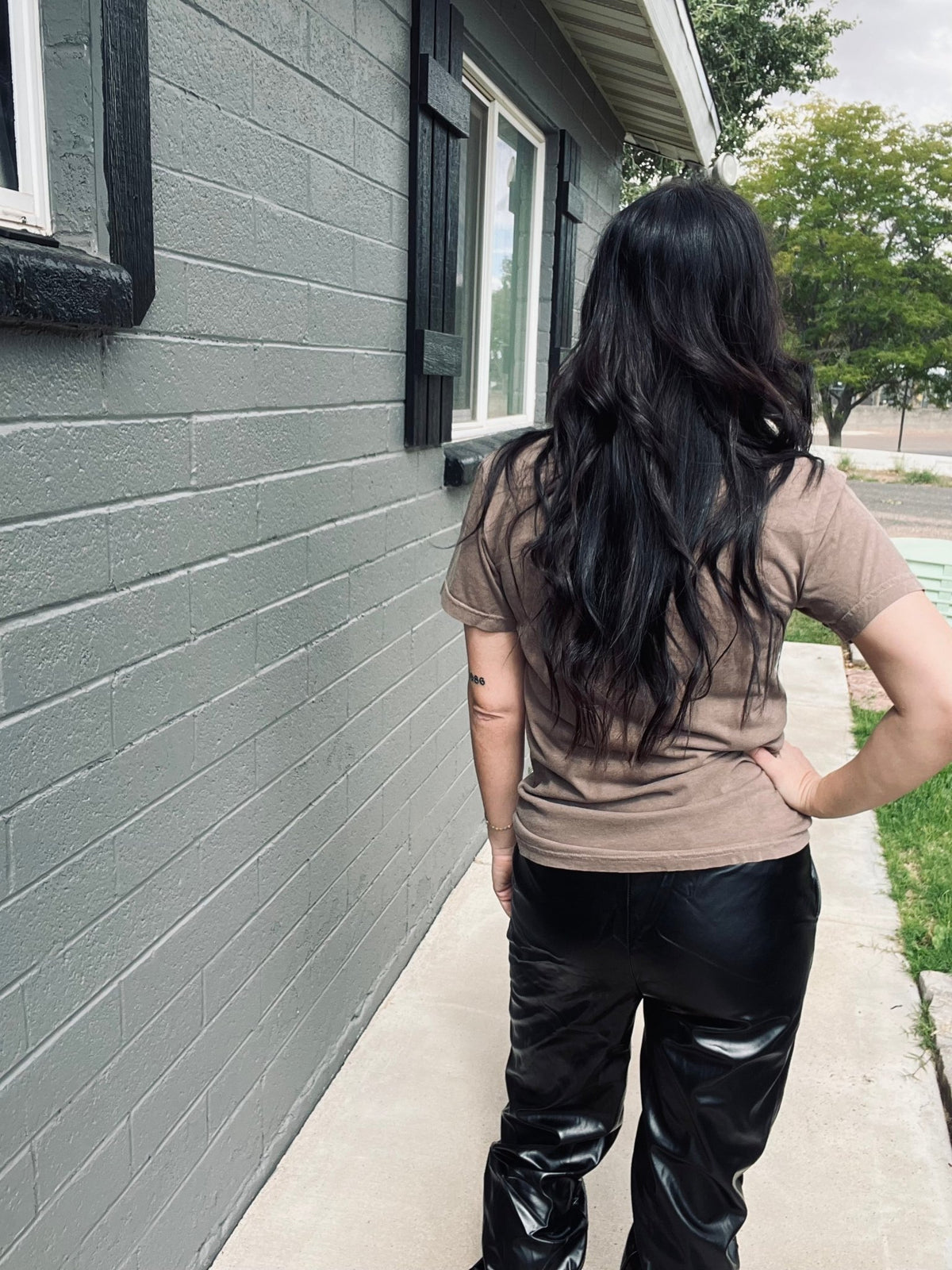 Person wearing a brown shirt and black leather pants standing on a sidewalk next to a gray house.