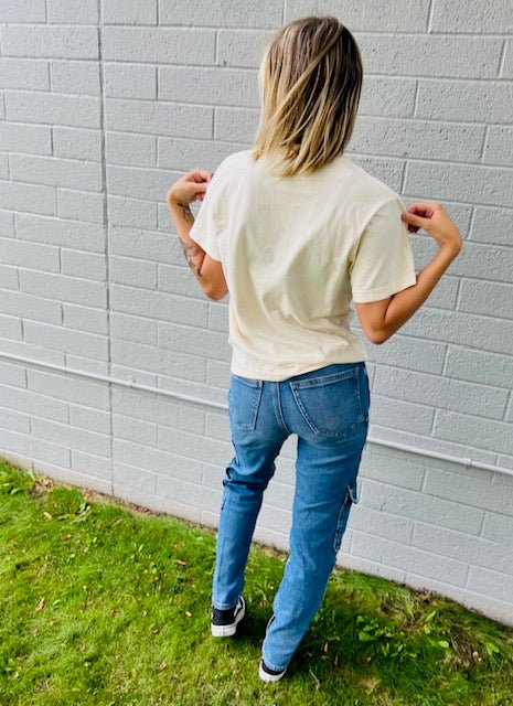 Person wearing a beige t-shirt and blue jeans standing against a gray brick wall.