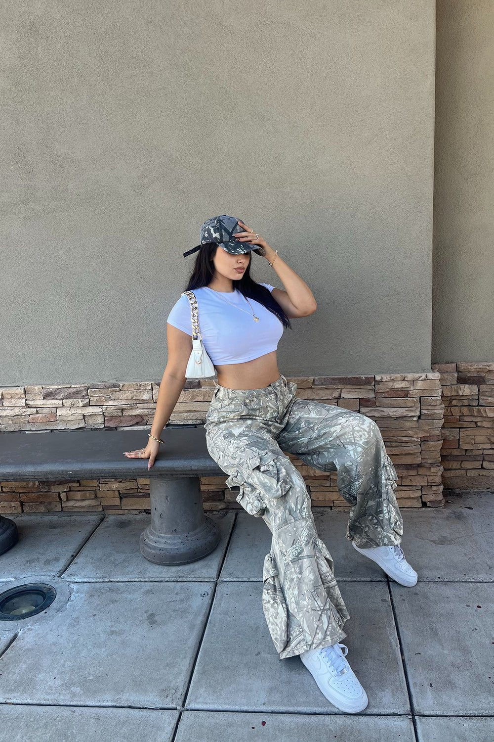 Woman sitting on a bench wearing a crop top, camouflage pants, and white sneakers.