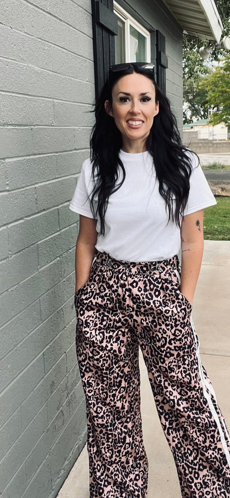 Woman wearing a white t-shirt and leopard print pants standing against a gray wall.