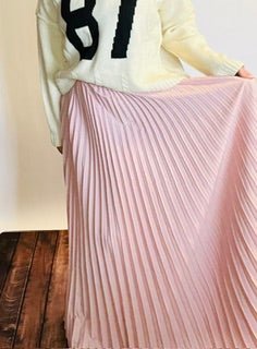 Person wearing a light-colored sweater and pink pleated skirt on a wooden floor.
