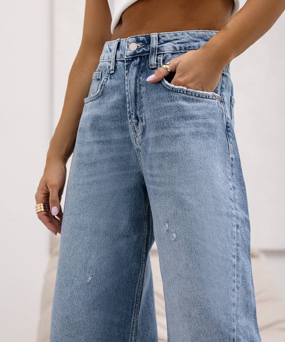 A close-up of a person wearing light-wash blue wide-leg denim jeans, with one hand in the pocket and the other resting at the side. The background is a soft, neutral indoor setting.