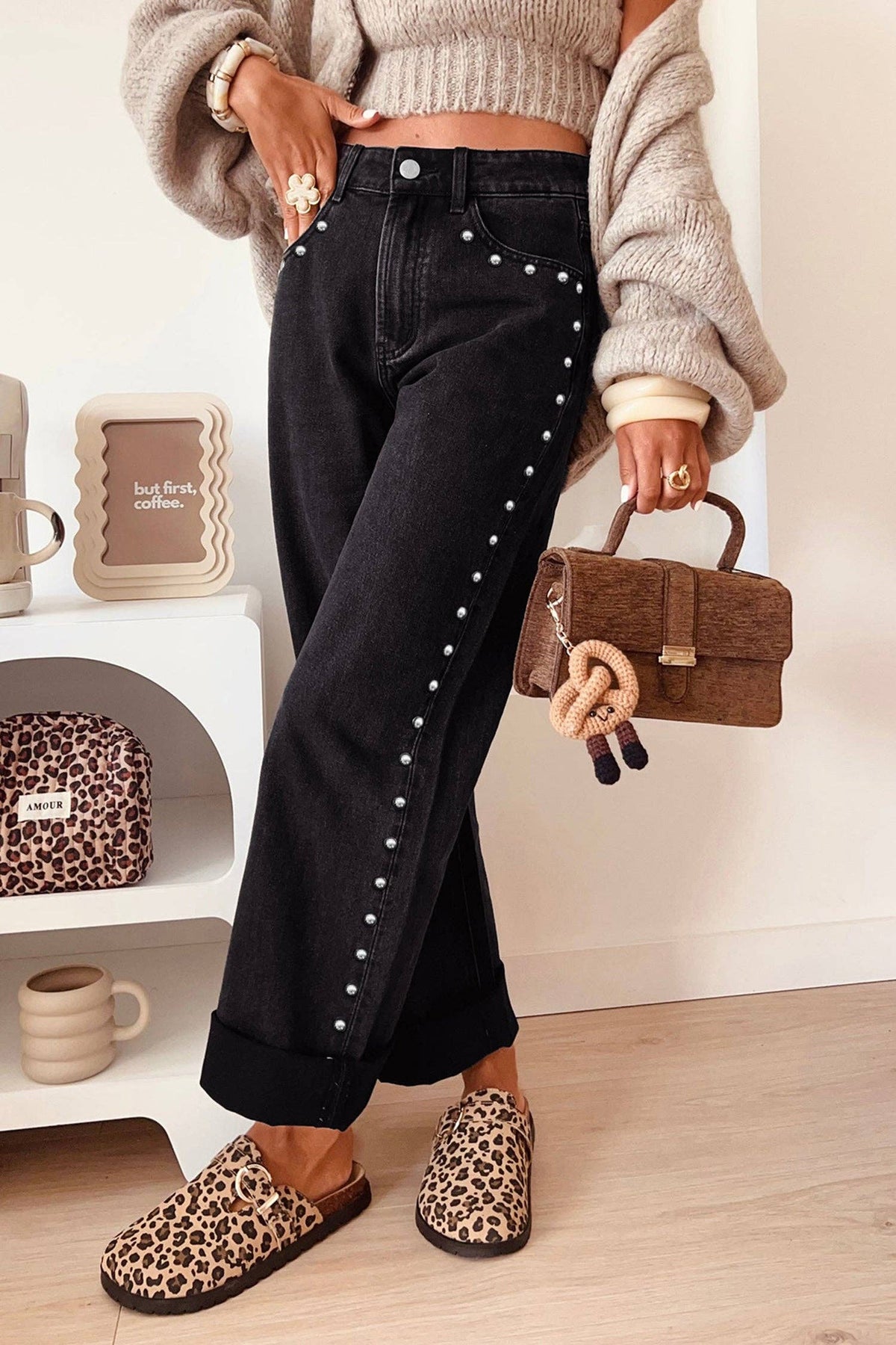 A person wearing black wide-leg jeans with silver stud detailing down the side, paired with leopard-print slip-on shoes and holding a brown textured handbag in a bright indoor setting.