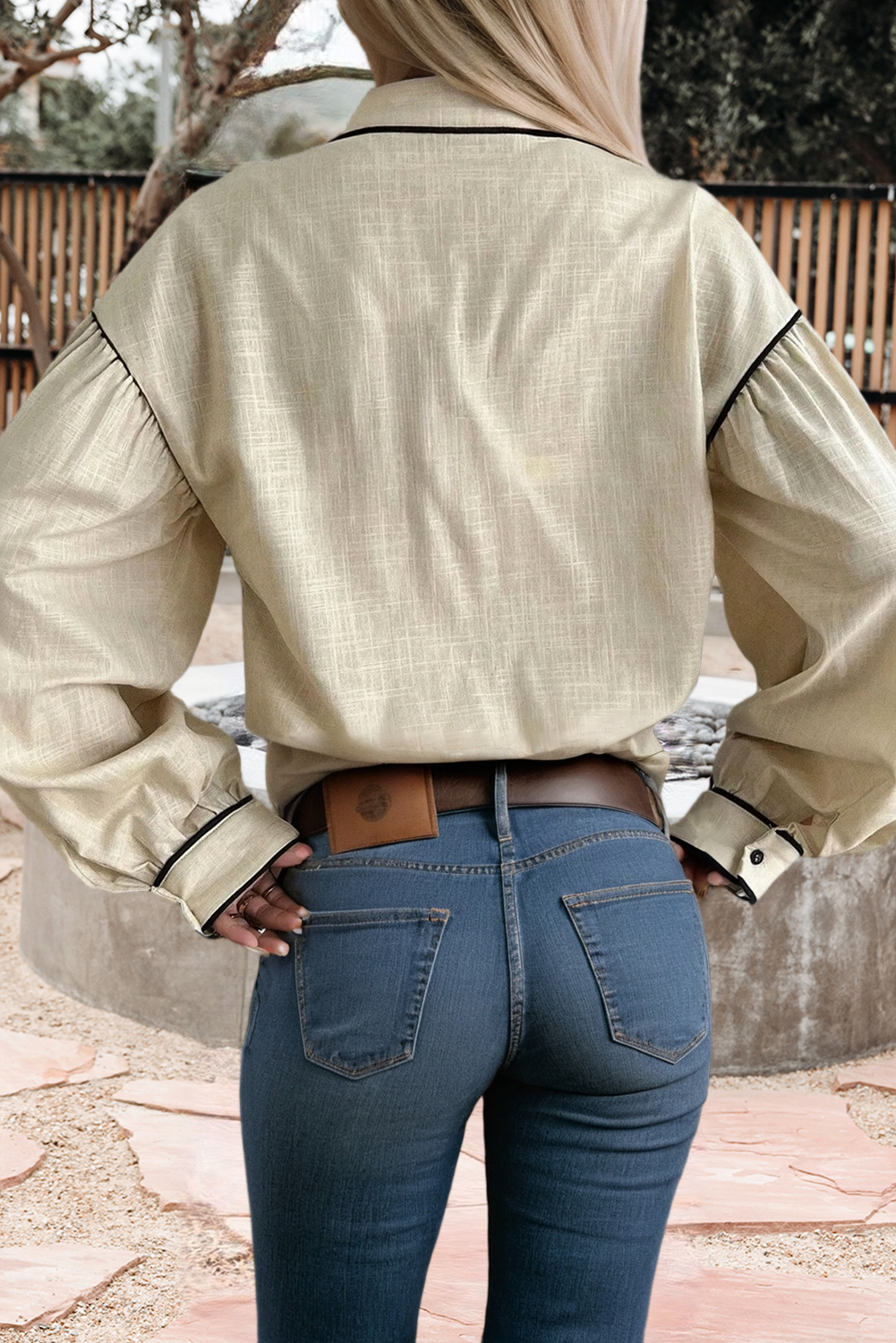 A woman viewed from behind wearing a light beige long-sleeve blouse with black piping details and blue jeans, standing outdoors.