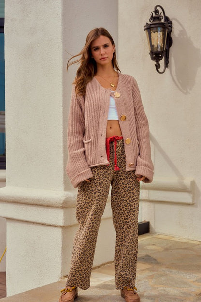 Woman wearing a pink cardigan and leopard print pants standing against a white wall.