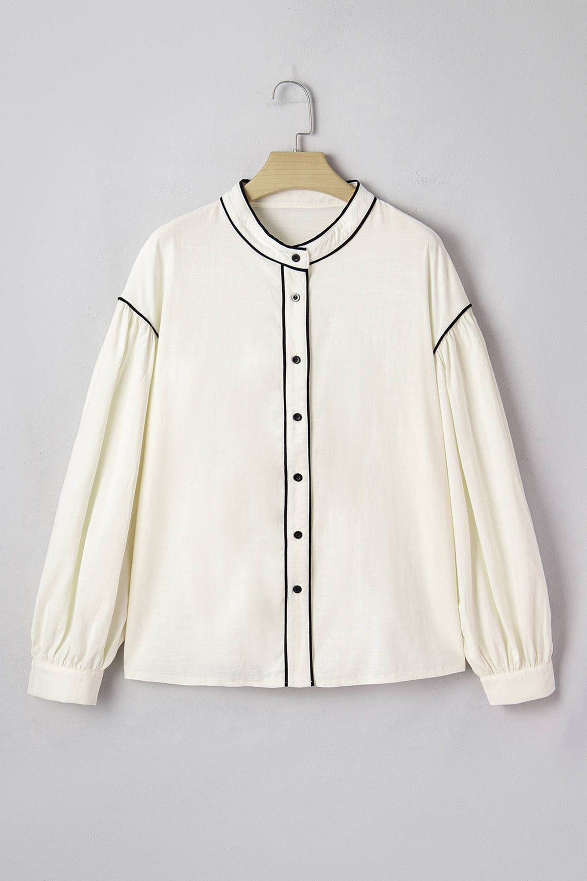 An off-white long-sleeve blouse with black piping trim and a button-up front, hanging on a wooden hanger against a light gray background.