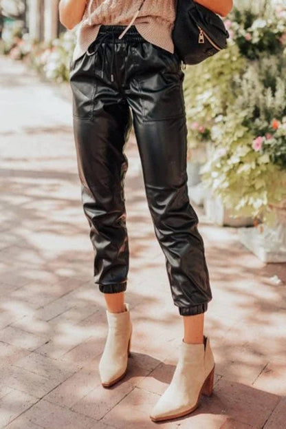 Person wearing black leather jogger pants and beige ankle boots on a sidewalk with flowers in the background