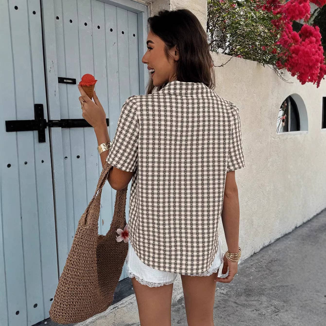 Woman in a checkered shirt and shorts eating ice cream, standing in front of a white door with a blue frame.