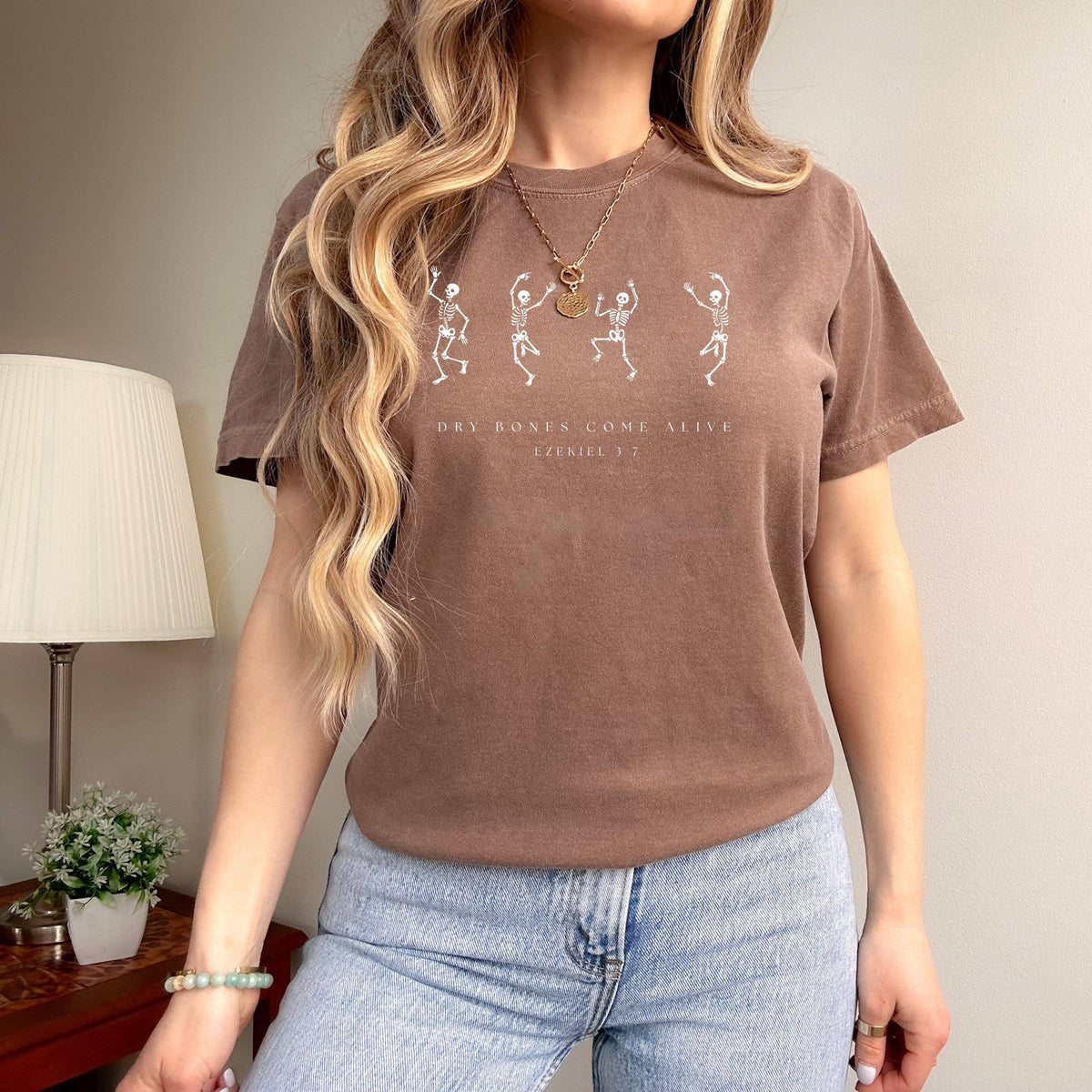 Person wearing a brown t-shirt with text and graphics, standing in a room with a lamp and plant.