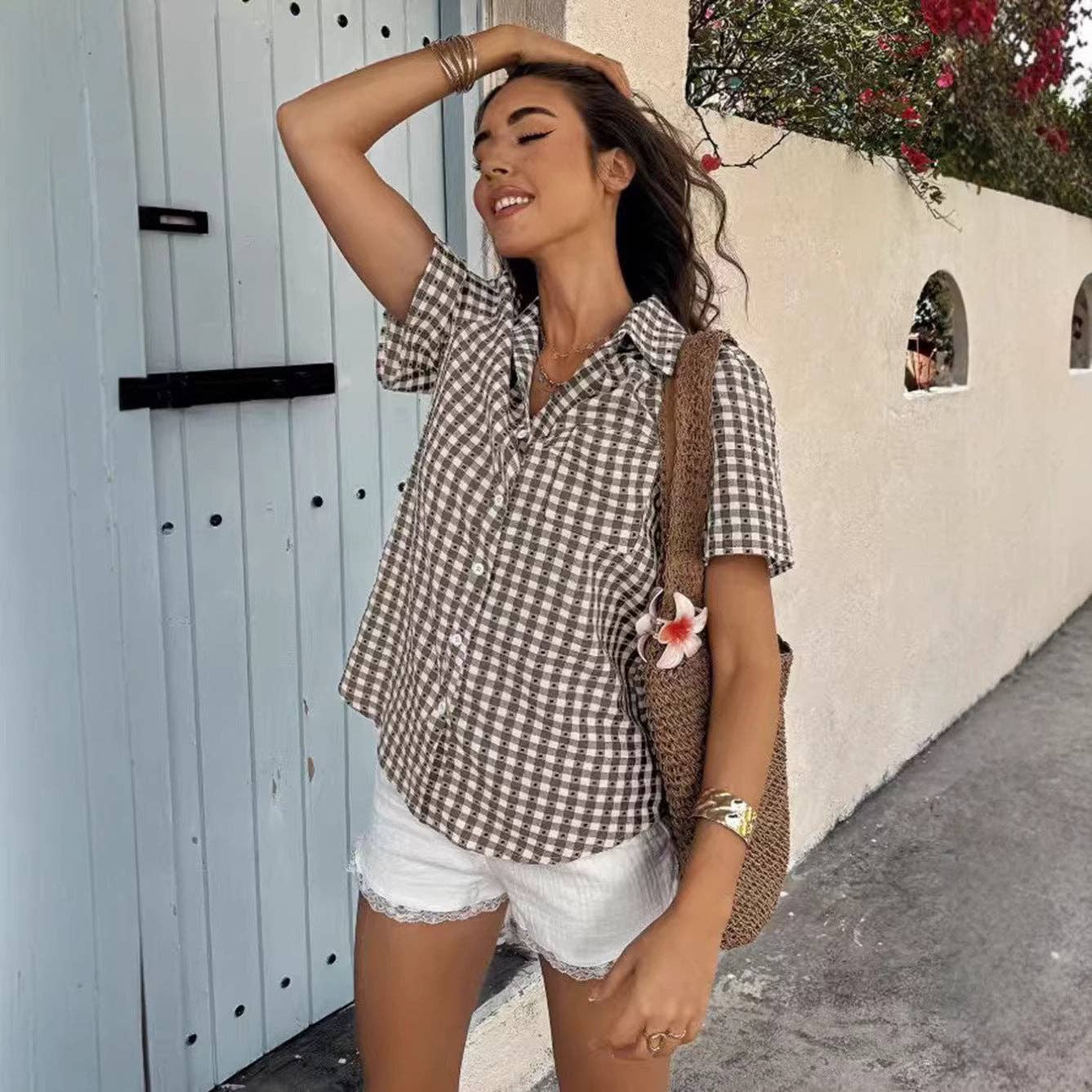 Woman in a checkered shirt and white shorts standing in front of a white wall with a blue door.