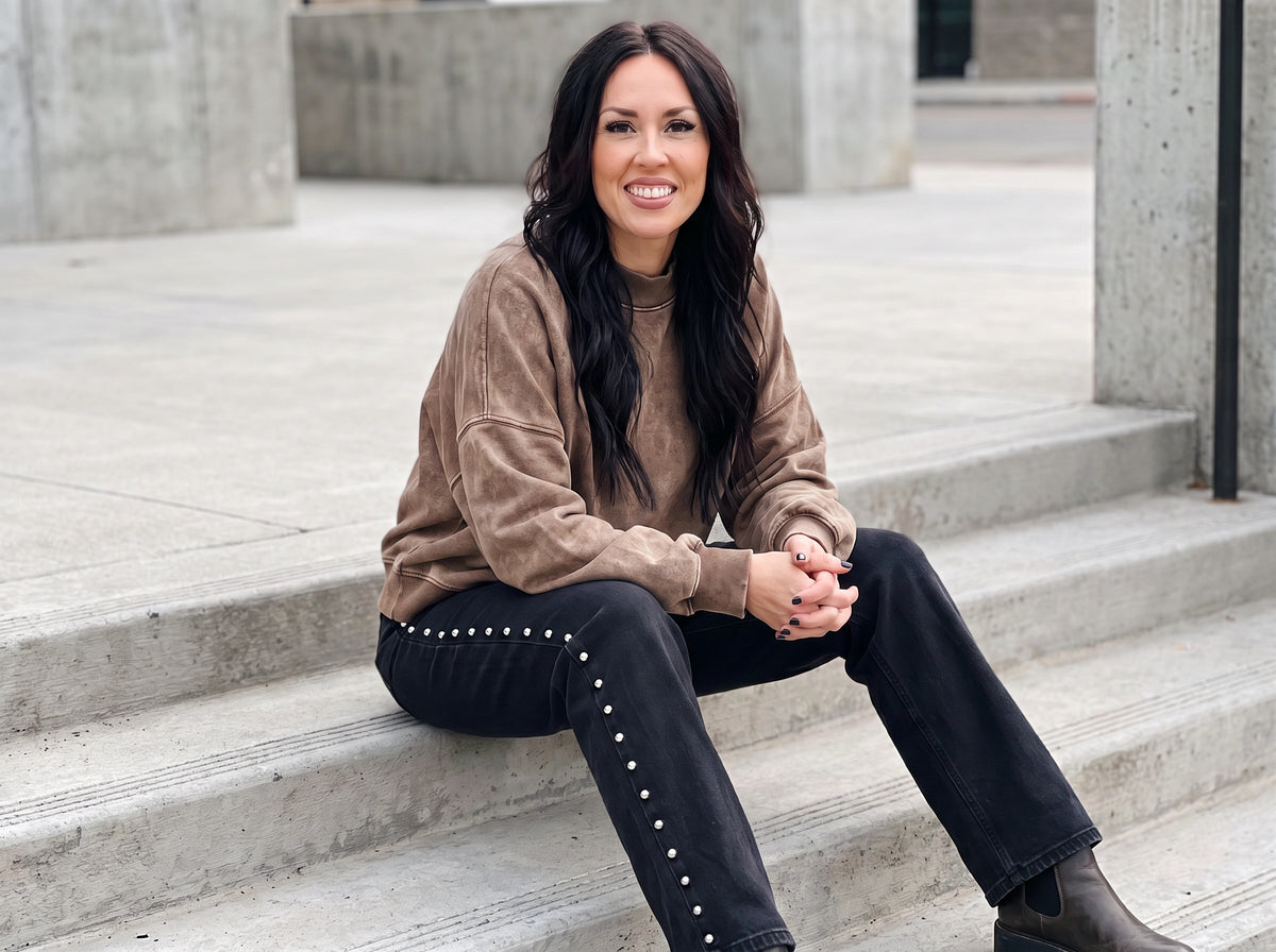 A woman with long dark hair sits on concrete steps wearing a brown long-sleeve top, black jeans with silver stud detailing, and black boots.