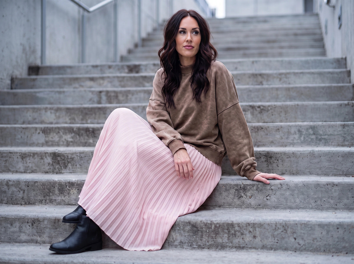 A woman sits on concrete stairs wearing a brown crewneck sweatshirt, a long light pink pleated skirt, and black ankle boots.