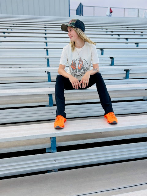 Person sitting on bleachers wearing a graphic t-shirt, black pants, and orange shoes.