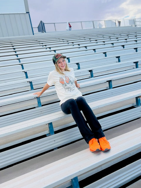 Person sitting on bleachers wearing a white t-shirt, dark jeans, and orange shoes.
