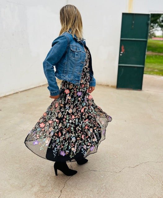 Person wearing a denim jacket and floral dress standing on a concrete surface with a white wall and green door in the background.