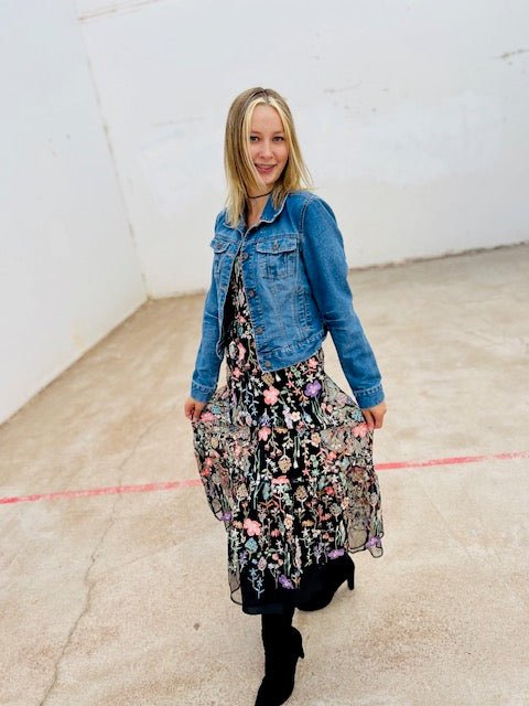 Woman wearing a floral dress and denim jacket in an indoor setting with a white wall.