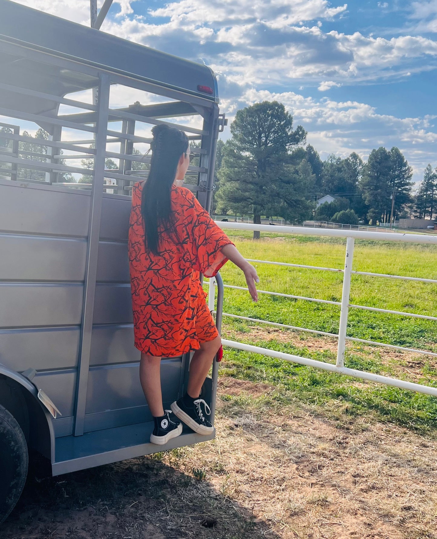 Person in an orange dress standing next to a livestock trailer in a rural setting with trees and open field.