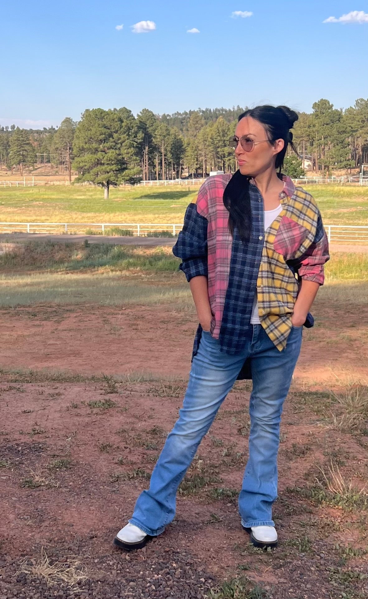 Person wearing a plaid shirt and jeans standing in an open field with trees in the background