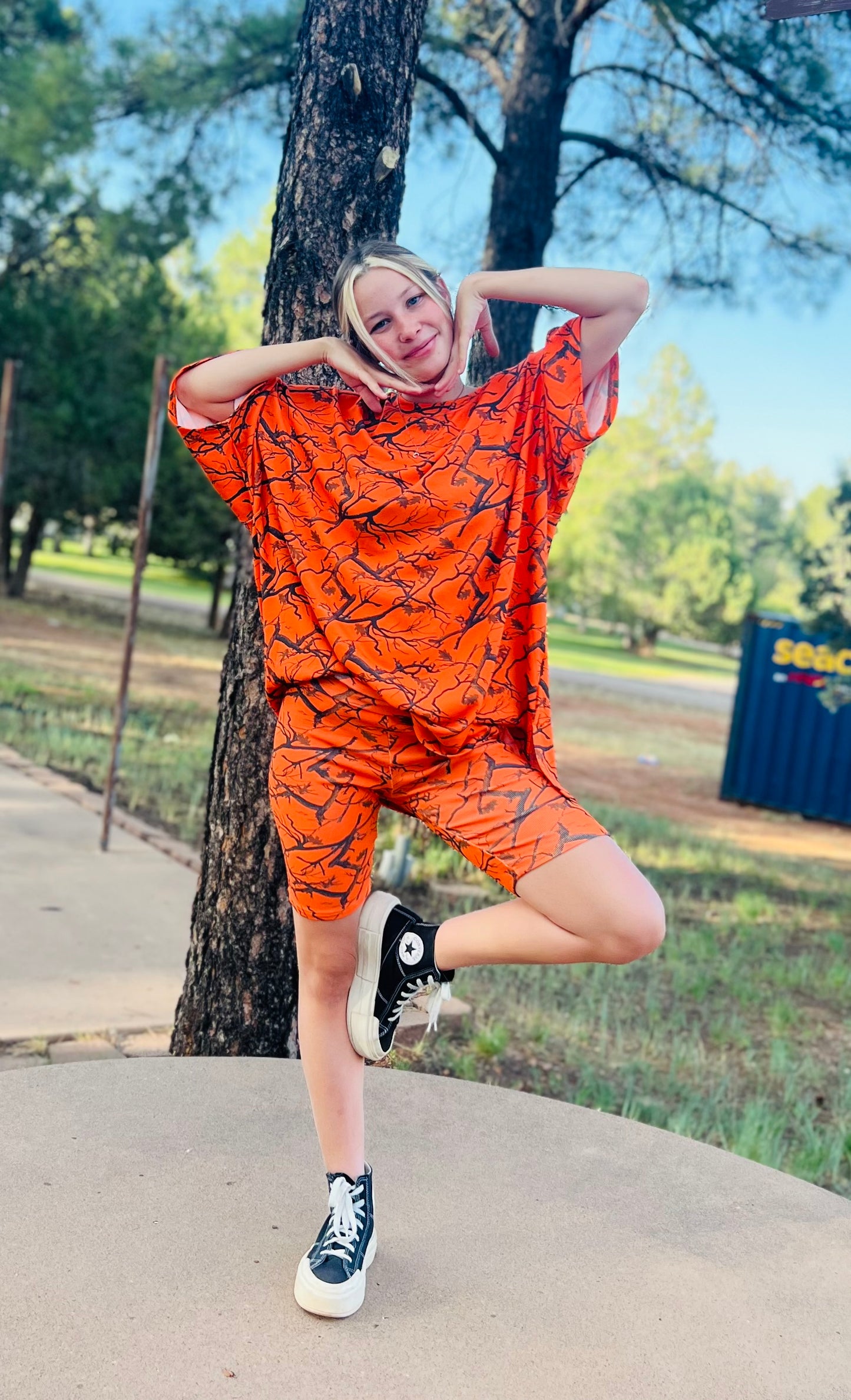 Person wearing an orange outfit with black patterns, standing on a sidewalk with trees and grass in the background.