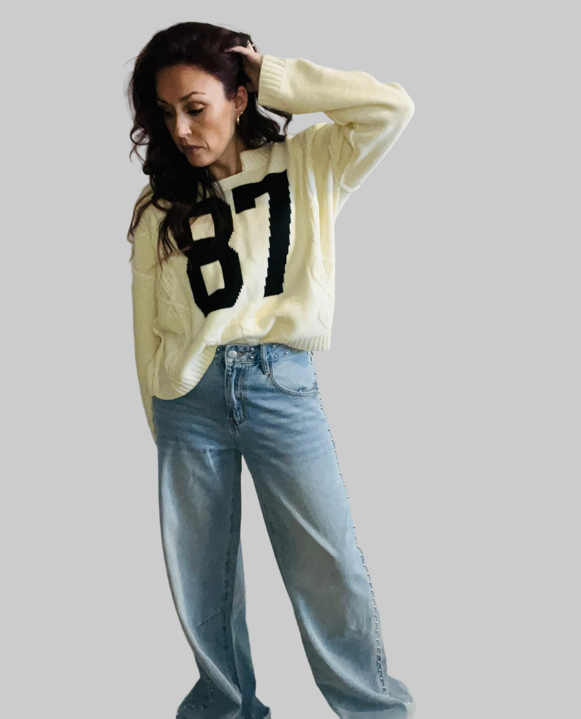 Woman wearing a light-colored sweater with a large number '87' and blue jeans on a gray background