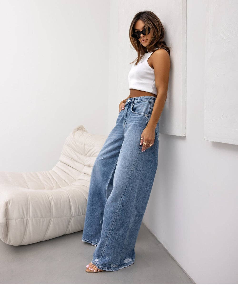A woman wearing a white sleeveless crop top and high-waisted, wide-leg light blue denim jeans stands in a minimalist white room next to a cream-colored lounge chair.