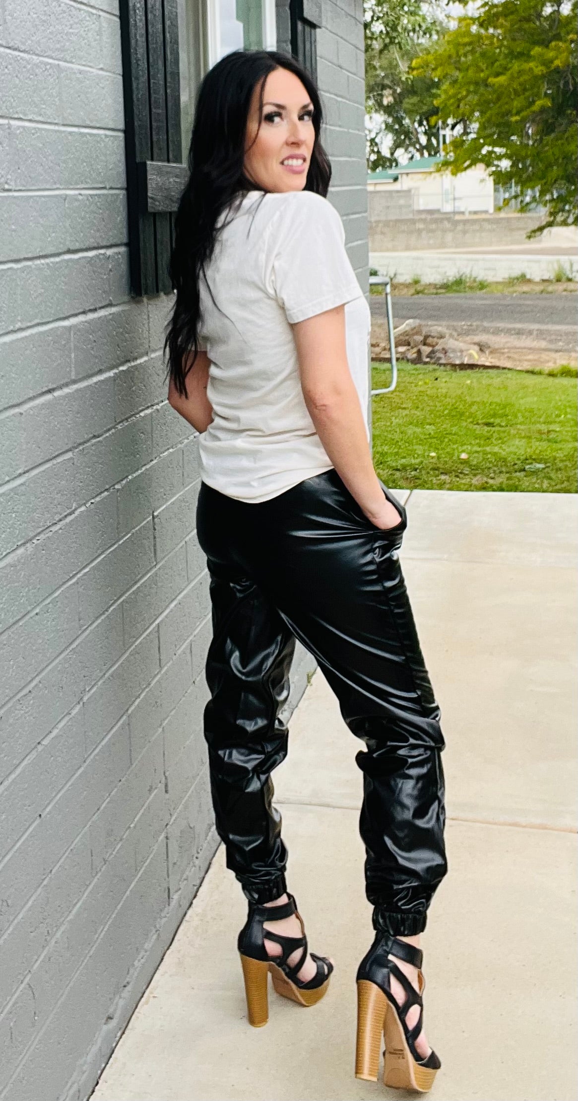 Woman wearing a white shirt and black leather pants leaning against a gray wall.