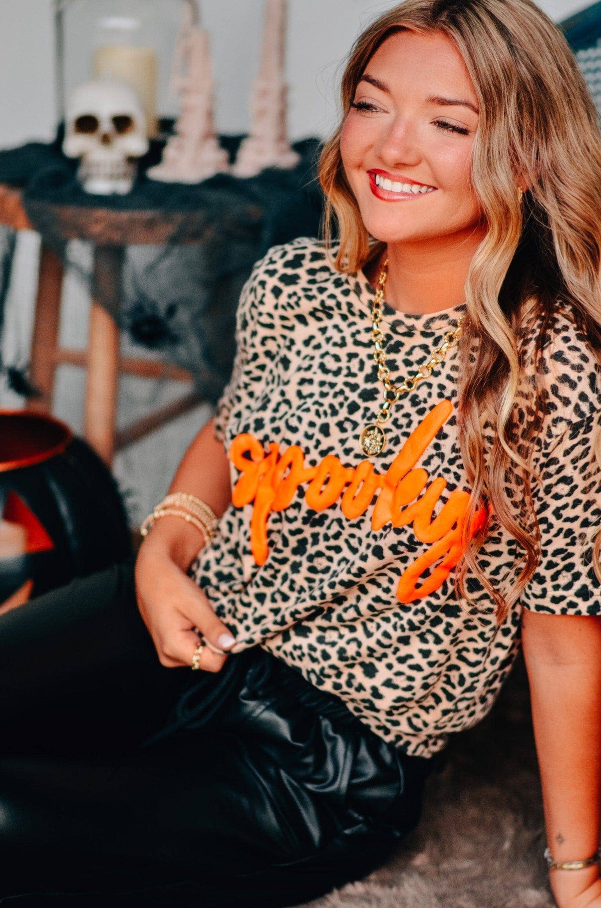 Woman wearing a leopard print shirt with orange text, sitting in a room with decorative items.