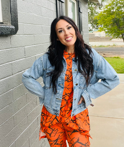Woman wearing an orange and black patterned outfit with a denim jacket standing against a gray brick wall.