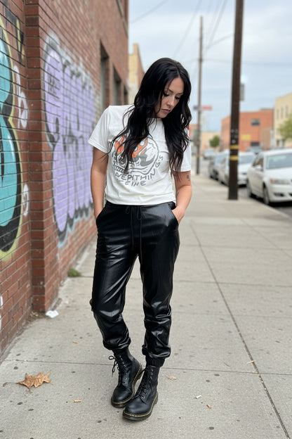 Person wearing a white graphic t-shirt, black leather pants, and black boots standing against a gray brick wall.