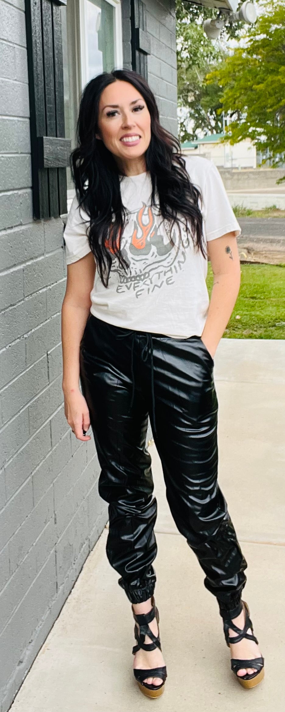 Woman wearing a graphic t-shirt and black pants standing against a gray wall.