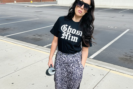 Woman wearing a black t-shirt with 'Ghost Him' text and patterned pants against a gray wall.