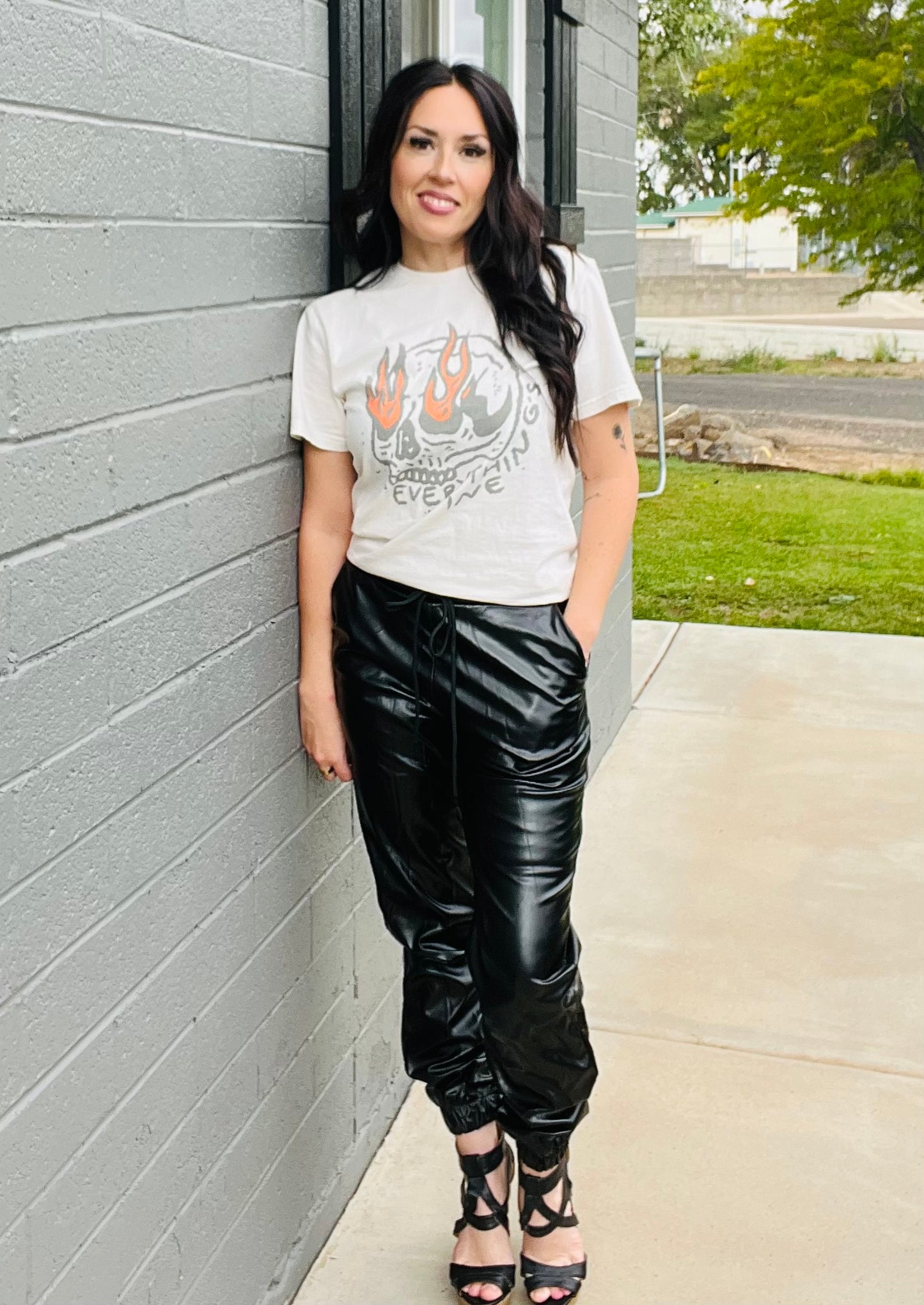 Woman wearing a graphic t-shirt and black pants leaning against a gray wall.