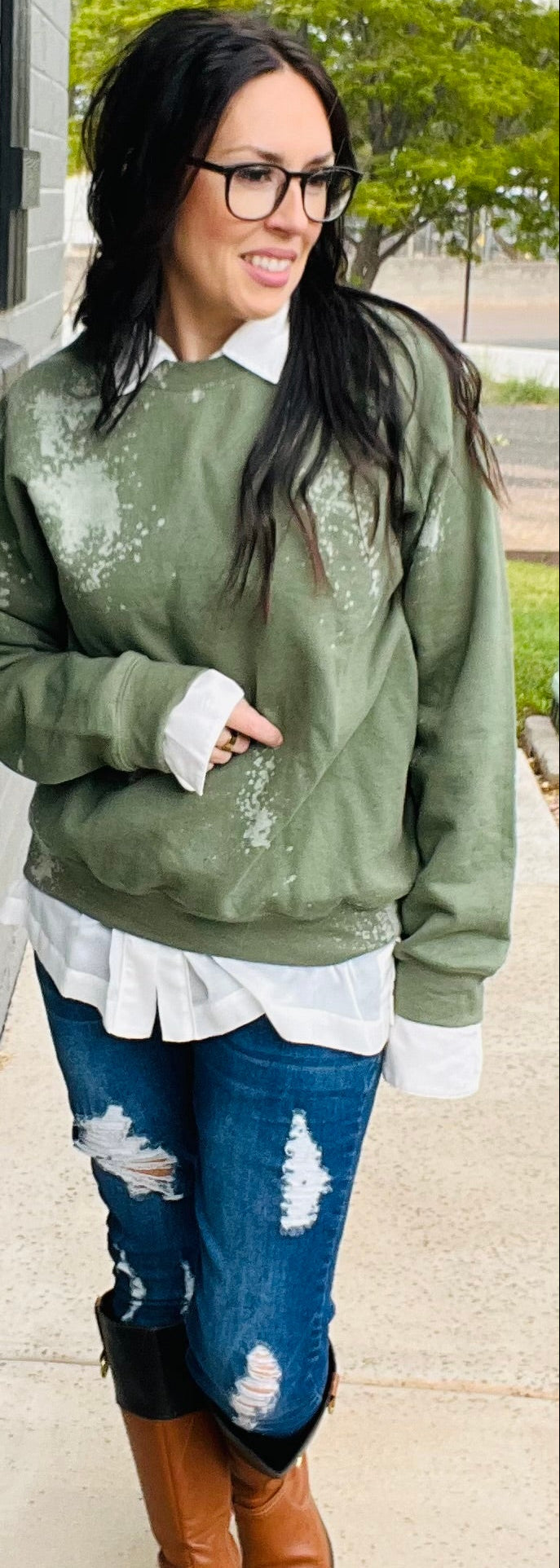Woman wearing a green distressed sweater, white shirt, blue jeans, and brown boots standing against a gray wall.