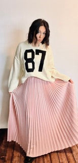 Person wearing a white sweater with black number 87 and a pink pleated skirt on a plain background