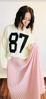 Woman wearing a white sweater with the number 87 and a pink skirt indoors.