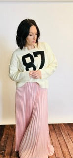 Woman wearing a white sweater with black numbers and pink pleated skirt indoors.