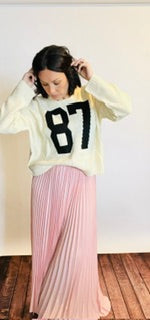 Person wearing a white sweater with black numbers and a pink pleated skirt indoors.