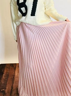 Person wearing a light-colored sweater and pink pleated skirt on a wooden floor.