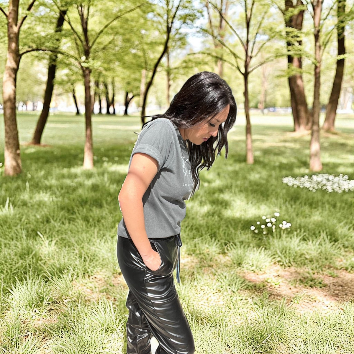 Woman in gray shirt and black pants standing in a park with trees and grass.
